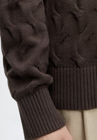 Manche de pull tricoté marron foncé avec bord-côte, montrant un motif torsadé texturé et une partie d’un pantalon beige clair.