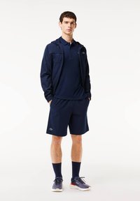 Navy athletic outfit featuring a hooded jacket, polo shirt, and shorts. Material appears lightweight. Includes green logo detail on shorts.