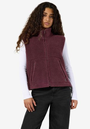 Vrouw met lang krullend haar die een mouwloos paarse fleece-vest draagt over een wit shirt met lange mouwen en een zwarte broek, staand tegen een witte achtergrond.