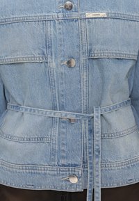 Veste en jean bleu clair avec des boutons argentés et une ceinture en jean assortie nouée à la taille, avec une petite étiquette "CLOSED" sur la poitrine.