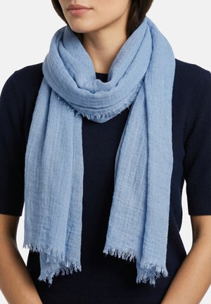 Woman wearing a loosely wrapped light blue fringed scarf over a short-sleeve navy sweater, facing front with neutral lips visible.