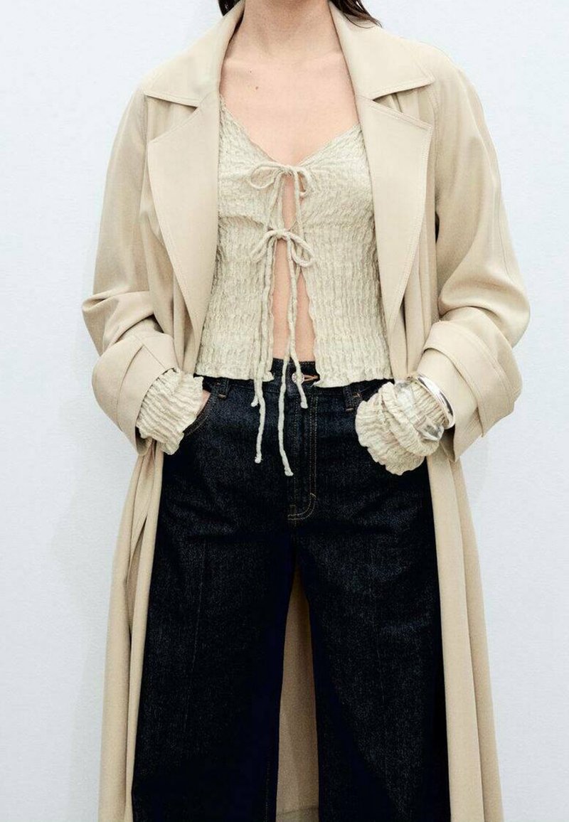 Femme portant un trench-coat beige par-dessus un top court texturé crème avec des liens, associé à un jean large taille haute de couleur sombre et mains dans les poches.