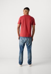 Camiseta de algodón roja con cuello redondo clásico, combinada con unos vaqueros de denim azul deslavado. Los vaqueros tienen un corte relajado y un desgaste sutil.