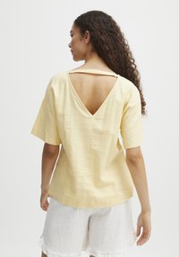 Blouse jaune à manches courtes avec un design en V profond au dos, fabriquée en tissu léger. Présente une coupe décontractée et des détails de couture simples.