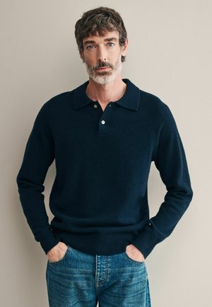 Navy knitted pullover with short collar, two button placket, and ribbed hem. Worn over light denim jeans, featuring a relaxed fit.