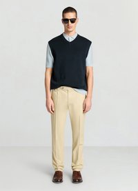 Pull sans manches bleu marine sur une chemise à col bleu ciel, associé à un pantalon beige et des chaussures marron. Des lunettes de soleil complètent le look.