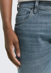 Hellblaue Jeans aus Denim mit glatter Textur, Fünf-Taschen-Design und dezentem Verblassen. Hand, die auf dem Oberschenkel aufliegt, zeigt die Passform.