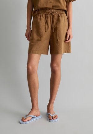 Personne portant un short marron ample avec des poches avant et une taille à cordon, associé à des tongs bleu clair sur un sol gris.