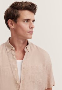 Kurzärmliges, hellbeiges Leinenhemd mit Button-Down-Kragen, Brusttasche und leicht strukturiertem Stoff. Über einem weißen Tanktop getragen.
