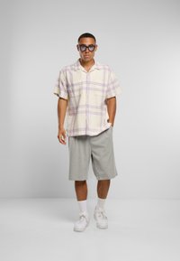 Camicia a maniche corte a quadri color crema con righe lavanda, abbinata a pantaloni corti grigi chiari fino al ginocchio e sneaker bianche. Occhiali da sole trasparenti indossati.