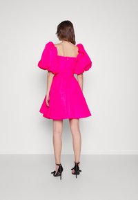 Vrouw die met de rug naar ons staat, gekleed in een felroze jurk met grote pofmouwen en zwarte hoge hakken, tegen een effen achtergrond.