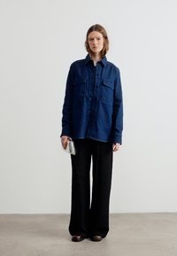 Γυναίκα που στέκεται φορώντας μπλε σκούρο πουκάμισο με κουμπιά, μαύρο παντελόνι φαρδύ, σκούρα παπούτσια και κρατώντας μικρό ασημένιο τσαντάκι.