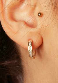 Złoty kolczyk w kształcie obręczy z fakturowaną powierzchnią, z rzędem przezroczystych kryształków wzdłuż zewnętrznej krawędzi. Noszony na płatku ucha.