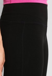 Czarne legginsy sportowe z wysokim pasem, o gładkiej fakturze i subtelnych detalach ściegów, w połączeniu z różową bluzką.