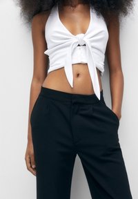 Haut blanc court avec un détail noué à l'avant, associé à un pantalon noir ajusté, présentant une texture lisse et une coupe droite.