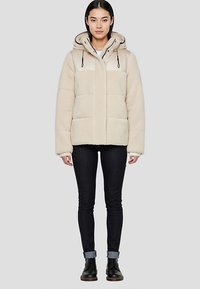 Veste en polaire beige avec capuche, dotée d'une fermeture à boutons, de grandes poches avant et de cordons élastiques. Portée avec un jean foncé et des bottes.