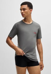 T-shirt gris à manches courtes avec un col rond et un logo "HUGO" rouge. Le tissu semble lisse et extensible. Le modèle porte un short noir.