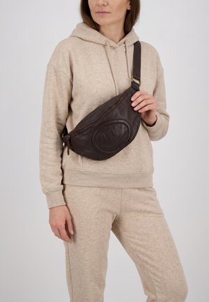 Vrouw draagt beige hoodie en broek met een donkerbruine leren crossbodytas met een geperst smiley gezicht.