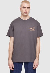Grå ribbet t-shirt med korte ærmer, der har et "Surf Club California" grafik i orange, båret med blå jeans.