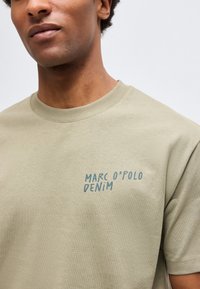 Κοντινό ενός ατόμου που φοράει μπεζ t-shirt με μπλε επιγραφή "MARC O'POLO DENIM" στο αριστερό στήθος.