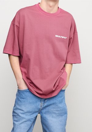 Personne portant un T-shirt ample couleur mauve avec l’inscription "MULTIPLY" sur la poitrine et un jean bleu clair, mains dans les poches, sur fond uni.