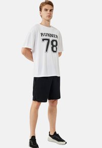 Hvid kortærmet skjorte med "RUNNER 78" i sort tekst, parret med sorte shorts. Modellen bærer sorte sportssko. Enkel, sporty design.