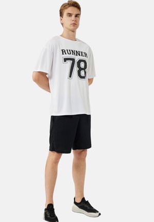 Camisa blanca de manga corta con "RUNNER 78" en texto negro, combinada con pantalones cortos negros. El modelo lleva zapatillas deportivas negras. Diseño simple y deportivo.