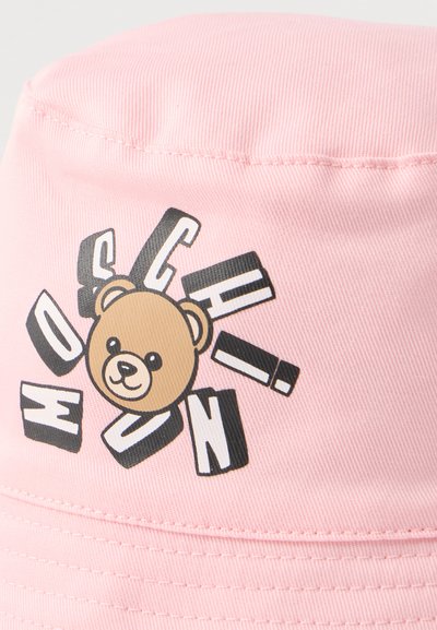 Ροζ καπέλο τύπου κουβά με πρόσωπο αρκουδάκι στο κέντρο, περιβαλλόμενο από διάσπαρτα μαύρα και λευκά γράμματα σε μπλοκ.