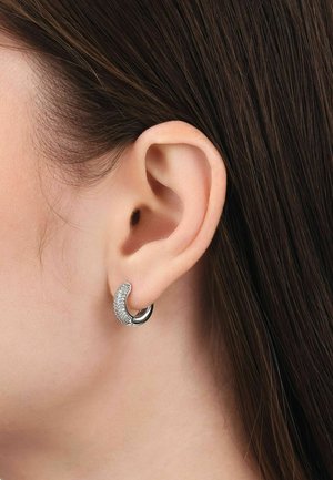 Primer plano de una oreja izquierda que lleva un pendiente de aro plateado parcialmente incrustado con pequeñas piedras preciosas claras, con cabello castaño liso visible.