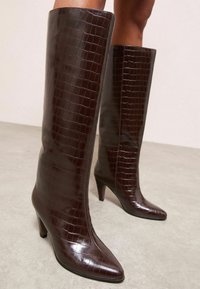 Rudos, blizgūs aukšti batai, pagaminti iš krokodilų odos imitacijos, su smailiu nosiu ir kelio ilgio dizainu. Lygi tekstūra su elegantišku paviršiumi.