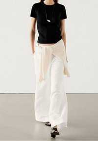 Maglietta nera aderente, pantaloni bianchi a gamba larga, cardigan beige chiaro legato in vita. Sandali neri con cinturini, collana di argento minimal.