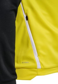 Chaqueta negra y amarilla con tejido suave, detalles en blanco, cremallera lateral y logo "BECOOL" cerca del dobladillo en gris.