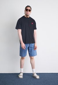Black cotton t-shirt with red heart logo, paired with light blue denim shorts, white crew socks, and grey sandals on a plain background.