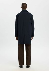 Marineblauer Überrock mit hohem Kragen, Rückenschlitz und langen Ärmeln, kombiniert mit braunen weit geschnittenen Hosen und schwarzen Schuhen. Strukturiertes Stoffdetail.