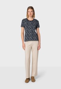 Korte mouwen marineblauwe t-shirt met een bloemenpatroon in wit en lichtblauw. Gecombineerd met crèmekleurige broeken en bruine sneakers, staand tegen een neutrale achtergrond.