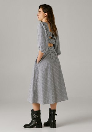 Naine kannab keskmise pikkusega triibulist kleiti tagaküljel lõigetega, kolmeveerlisi varrukaid, käed taskutes ja musti pahkluu saapaid pandlatega.