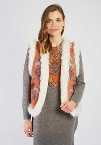 Brązowa kamizelka z białym sztucznym futrem i kolorowym haftem w kwiaty. Noszona na szarej dzianinowej sukience z żywym geometrycznym wzorem.