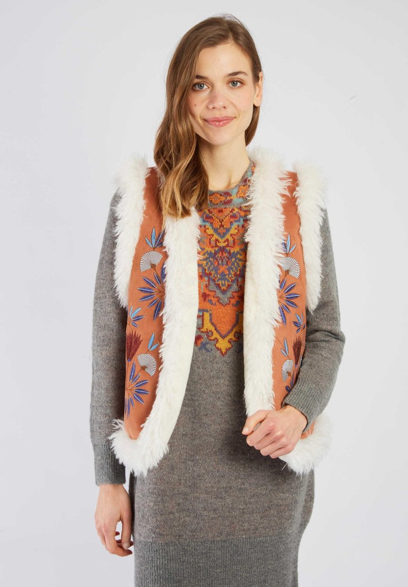 Brązowa kamizelka z białym sztucznym futrem i kolorowym haftem w kwiaty. Noszona na szarej dzianinowej sukience z żywym geometrycznym wzorem.