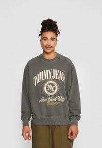 Felpa grigia con scollatura a girocollo e polsini a costine, con stampa "TOMMY JEANS" in giallo e un logo circolare etichettato "New York City."