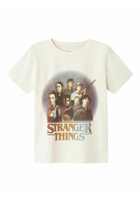 Off-white bomuld t-shirt med grafisk tryk af flere karakterer fra "Stranger Things" samt seriens titel i blå og sort.
