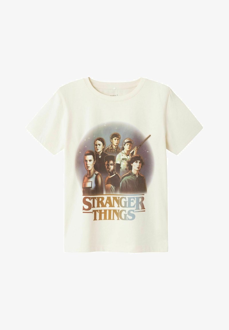 Bela bomba iz 100% bombaža v off-white barvi, ki vsebuje grafični tisk več likov iz serije "Stranger Things" ter naslov oddaje v modri in črni barvi.