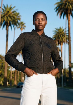 Femme aux cheveux courts portant une veste matelassée noire et un pantalon blanc, debout les mains dans les poches dans une rue bordée de palmiers.