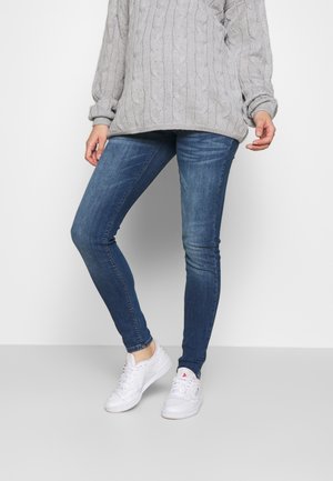 Person, die blaue Skinny-Jeans, weiße Sneakers und einen hellgrauen Zopfpullover trägt, steht vor einem schlichten Hintergrund.