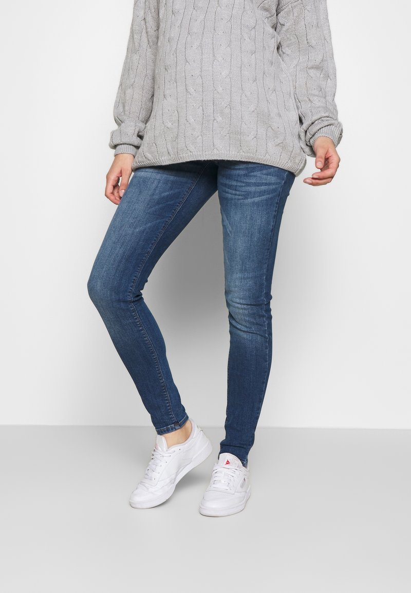 Personne portant un jean skinny bleu, des baskets blanches et un pull en maille torsadée gris clair, debout devant un fond uni.