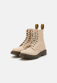 Stivaletti in pelle beige con punta rotonda, lacci sul davanti, cuciture gialle e linguette di aiuto con testo nero sopra. Suola in gomma testurizzata.