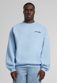 Gaiši zils pelēkpromaksts ar ribotiem piedurkņu un apakšmalas galiem, uz krūtīm ir neliela melna logo. Izgatavots no mīksta auduma ar brīvu piegriezumu.