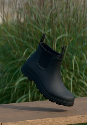 Zwarte rubberen enkellaars met een gladde afwerking en een gestructureerde zool, voorzien van een trekkoord voor gemakkelijk aan- en uittrekken, geplaatst op een houten oppervlak.