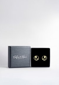 Guldtonede cirkulære øreringe i en sort fløjl displayboks, med et udskåret design af en figur indenfor en cirkel, sat mod en tekstureret sort kasse.