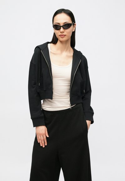 Sweatshirt preta curta com fecho de correr e bolsos frontais, usada por cima de uma camisola de alças bege claro. Calças pretas de perna larga completam o conjunto.