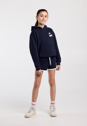 Jongere staand in een marineblauwe hoodie met een wit kersenlogo, marineblauwe korte broek met witte rand, witte sneakers en gestreepte sokken tegen een effen achtergrond.